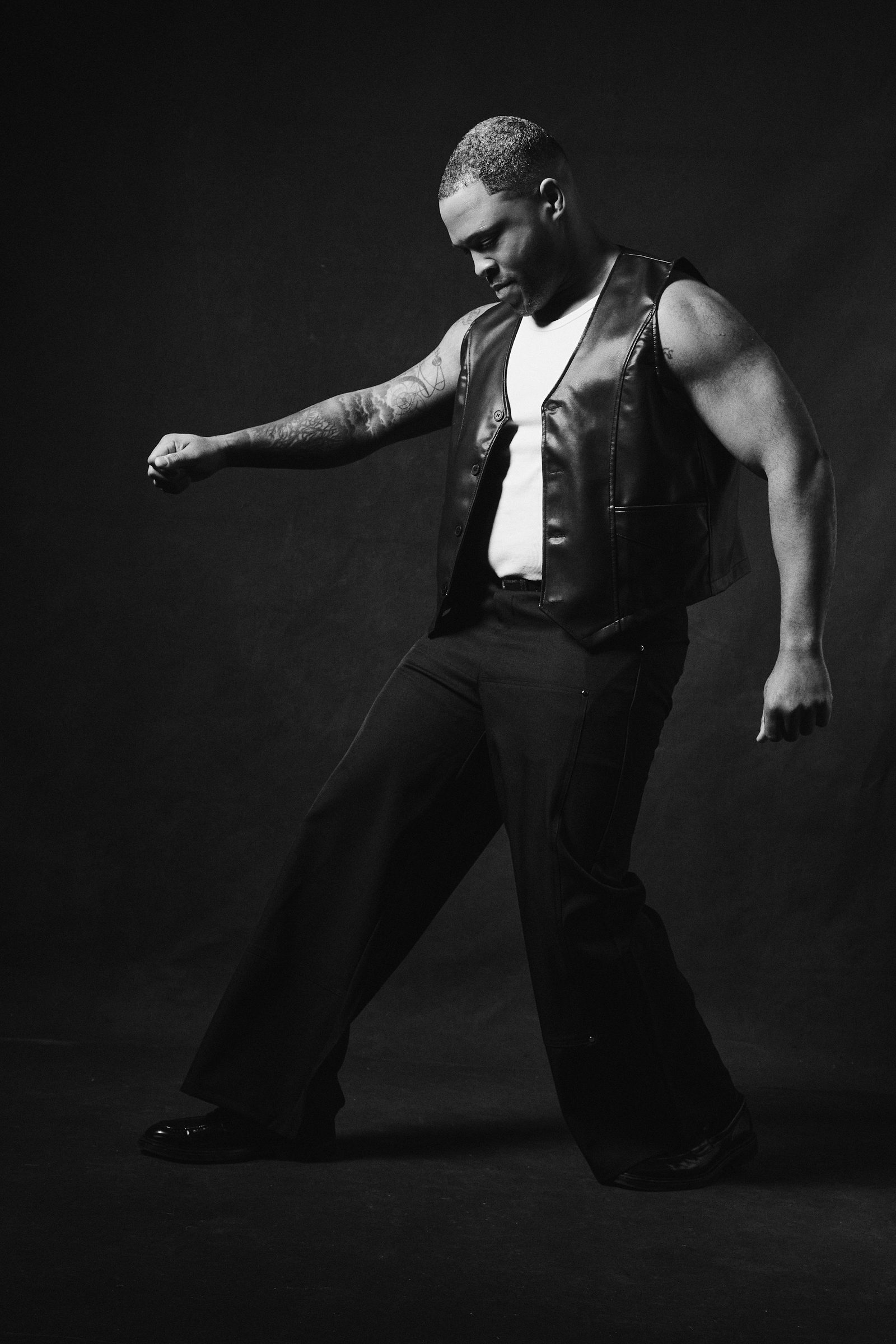 Dion Dawson mid-step in a black leather vest and white tank, black-and-white studio portrait.
